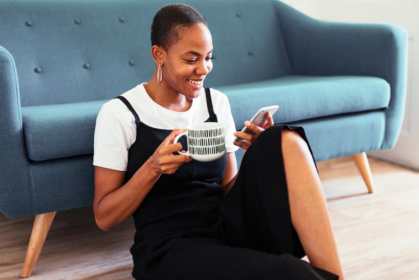 A woman sitting on the floor, drinking a cup of coffee, while looking at her phone