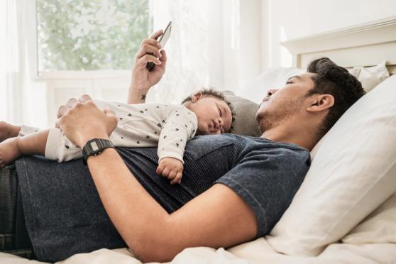 A man using his phone laying in bed with his infant child