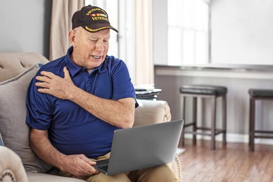 Senior man holding his shoulder while on the computer on the couch
